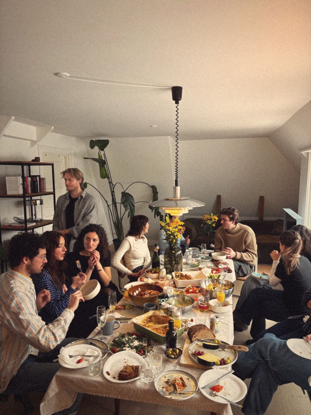 A long table in a bright attic apartment, covered in salads, bread and cheese, with a dozen guests talking