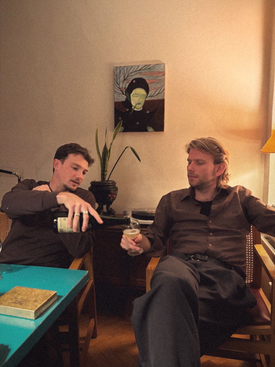 Two friends in armchairs, one pouring a glass of sparkling wine for the other