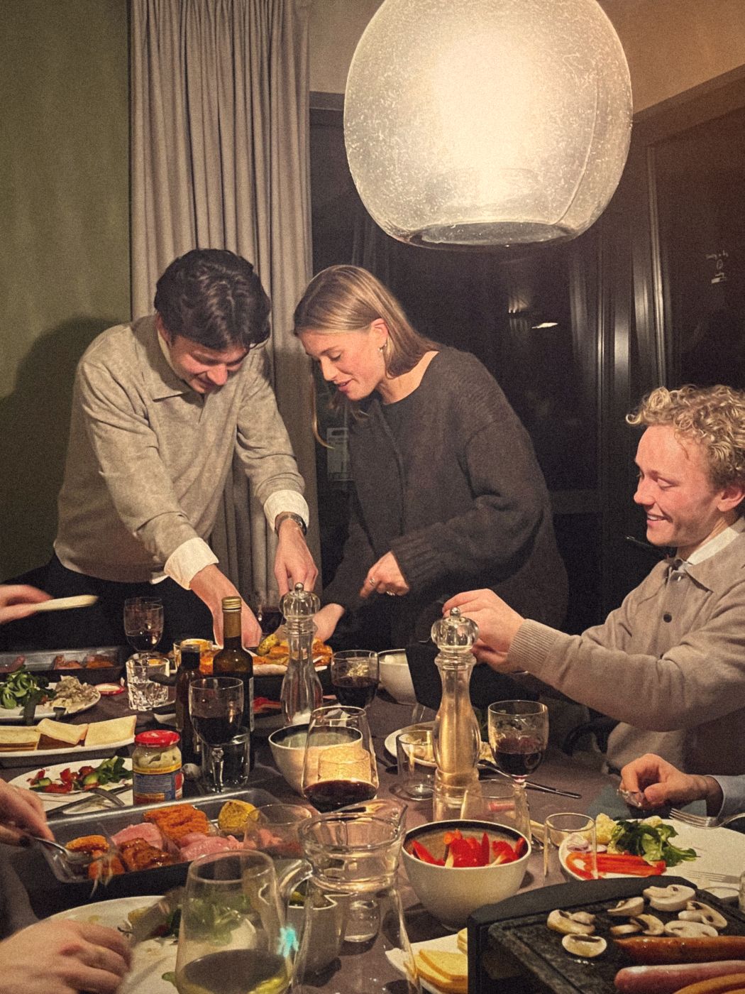 Friends leaning over a crowded dinner table under a glass lamp, sharing food
